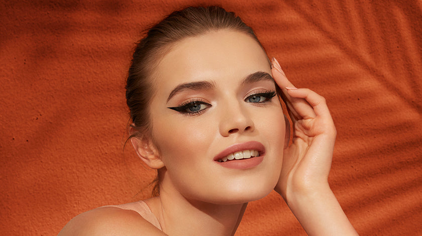 Model with bold winged eyeliner and a flawless makeup look, posing against a warm orange textured background.