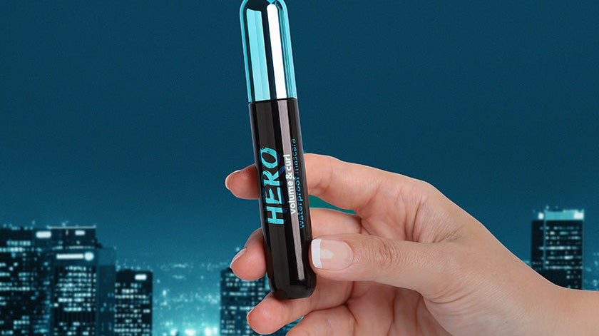 A hand holding Flormar Hero Waterproof Mascara against a nighttime cityscape. The mascara features a sleek black and blue design, emphasizing volume and curl for bold lashes.